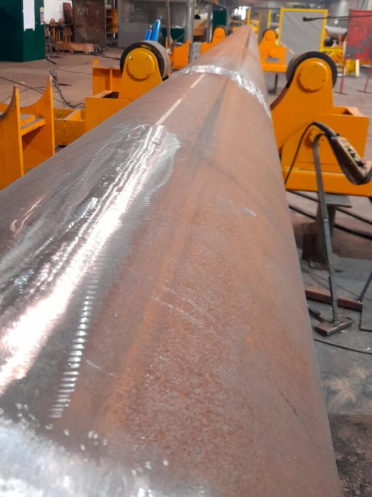 Una imagen que muestra un tubo metálico largo en un taller industrial, con maquinaria pesada y estructuras de metal alrededor.