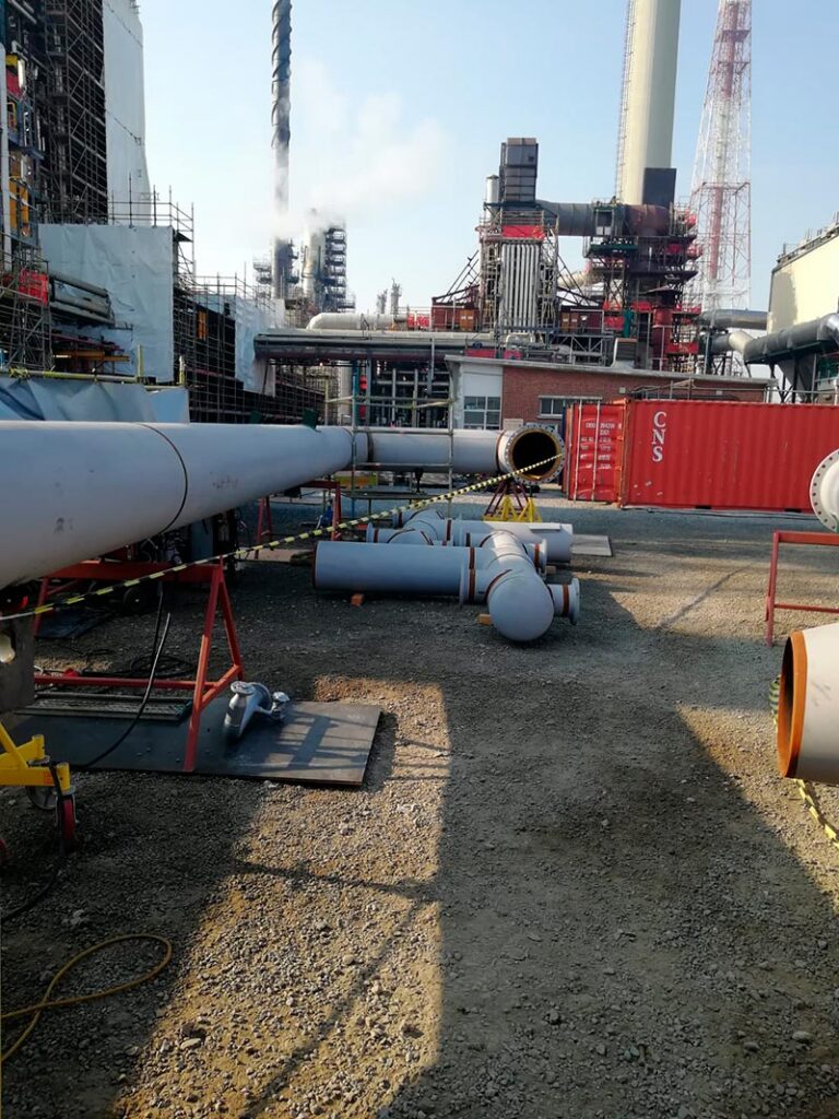 Fotografía de una planta industrial con varios tubos grandes y estructuras metálicas en primer plano y chimeneas emitiendo vapor en el fondo.